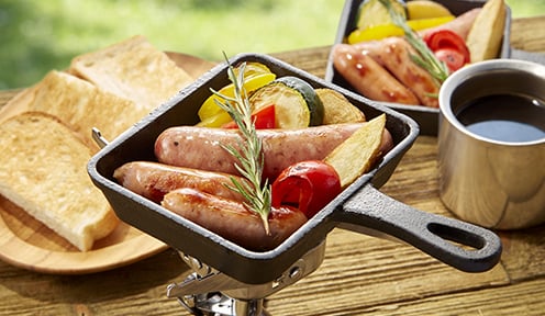 ガスバーナーの上に置いた四角いスキレットの中に良い焼き色のついたウインナーやじゃがいもパプリカなどの野菜のうえにハーブのような香草が乗せてある。その奥には焼いたトーストが数枚とコーヒーも置いてある食欲がそそられる写真